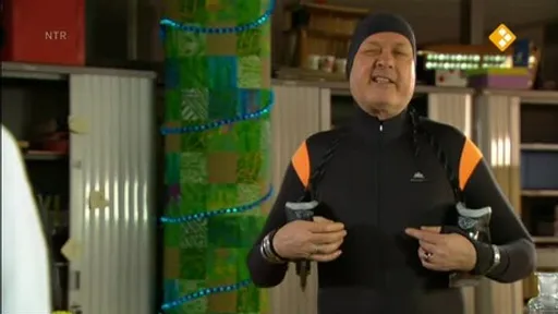 Persoon in zwart sportpak en muts houdt wandelstokken vast en glimlacht in een binnenruimte.