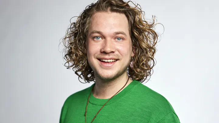 Glimlachende persoon/Pascal Tan met halflang krullend haar, groene trui, neuspiercing en lichte achtergrond.