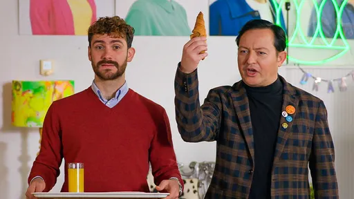 Twee mannen staan naast elkaar. Één heeft een dienblad vast met een glas met sap erop en de man ernaast houdt een croissant in de lucht.