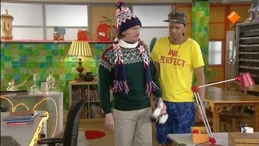Twee mannen in opvallende kleding staan naast elkaar in een kleurrijke kamer; één draagt een muts, de ander een geel T-shirt met tekst.