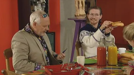 Man in beige colbert kijkt naar zijn telefoon terwijl een glimlachende man naast hem een broodje omhooghoudt aan een gedekte tafel.